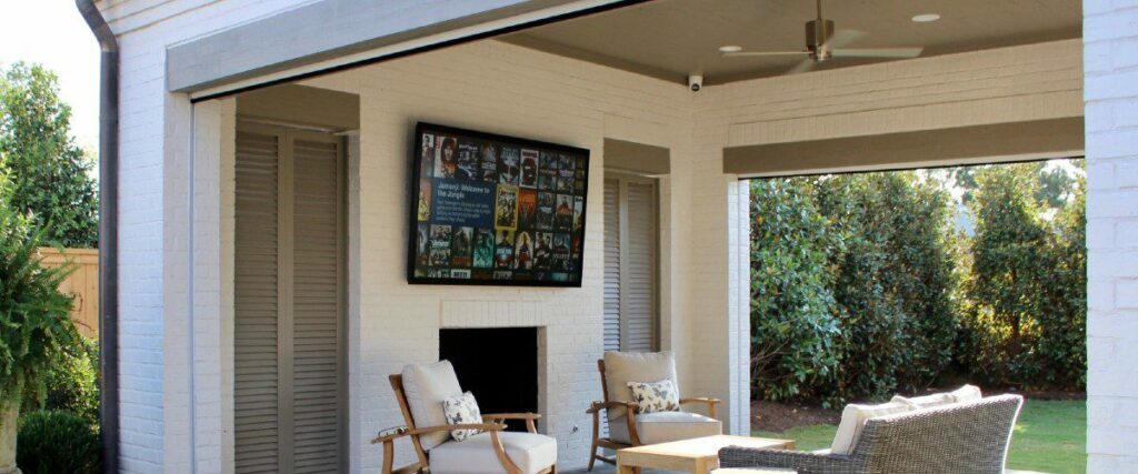 Outdoor TV mounted over fireplace in patio with visible painted white brickwork and bushes in the backgorund.